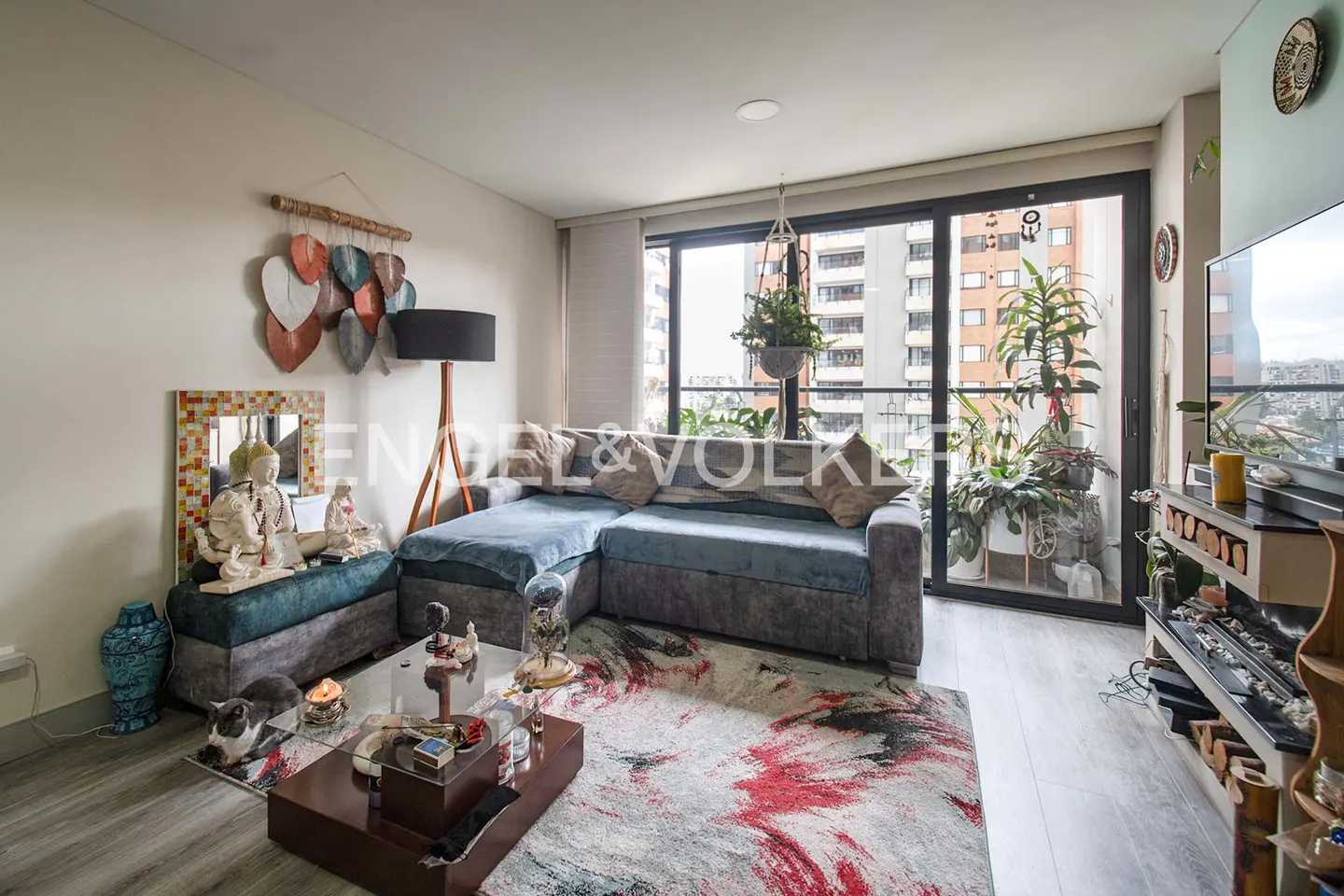 Living room with a blue sectional sofa, glass coffee table, and a colorful rug. Balcony with plants and city view. Buddha statues and wall art.