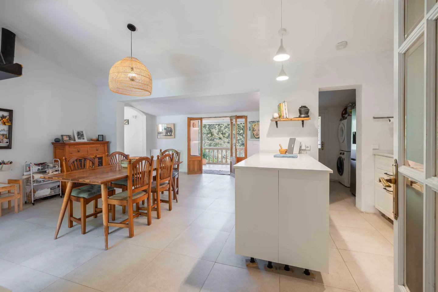 Bright, open-concept kitchen and dining area with white walls, tile floors, and wooden furniture. A woven pendant light hangs above the dining table.