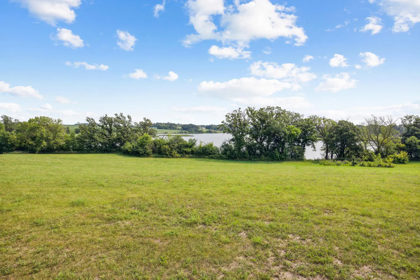 A grassy field leads to trees and a lake under a blue, cloudy sky.