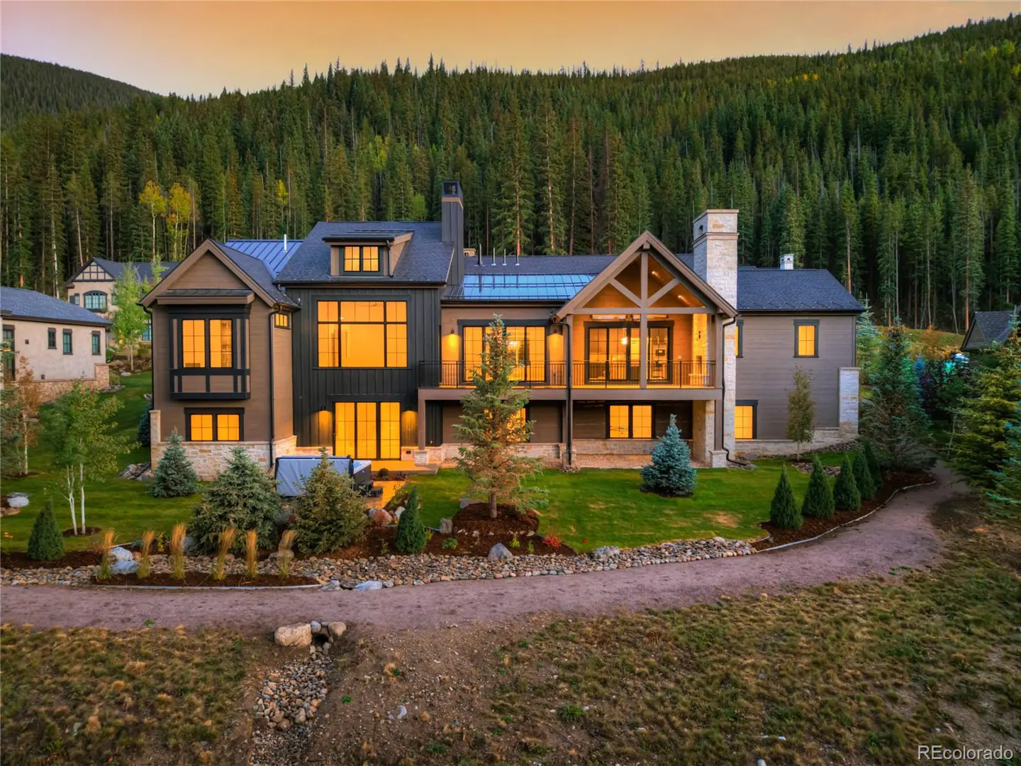 A large, modern mountain home with dark siding and many windows sits on a green lawn with a forest backdrop.
