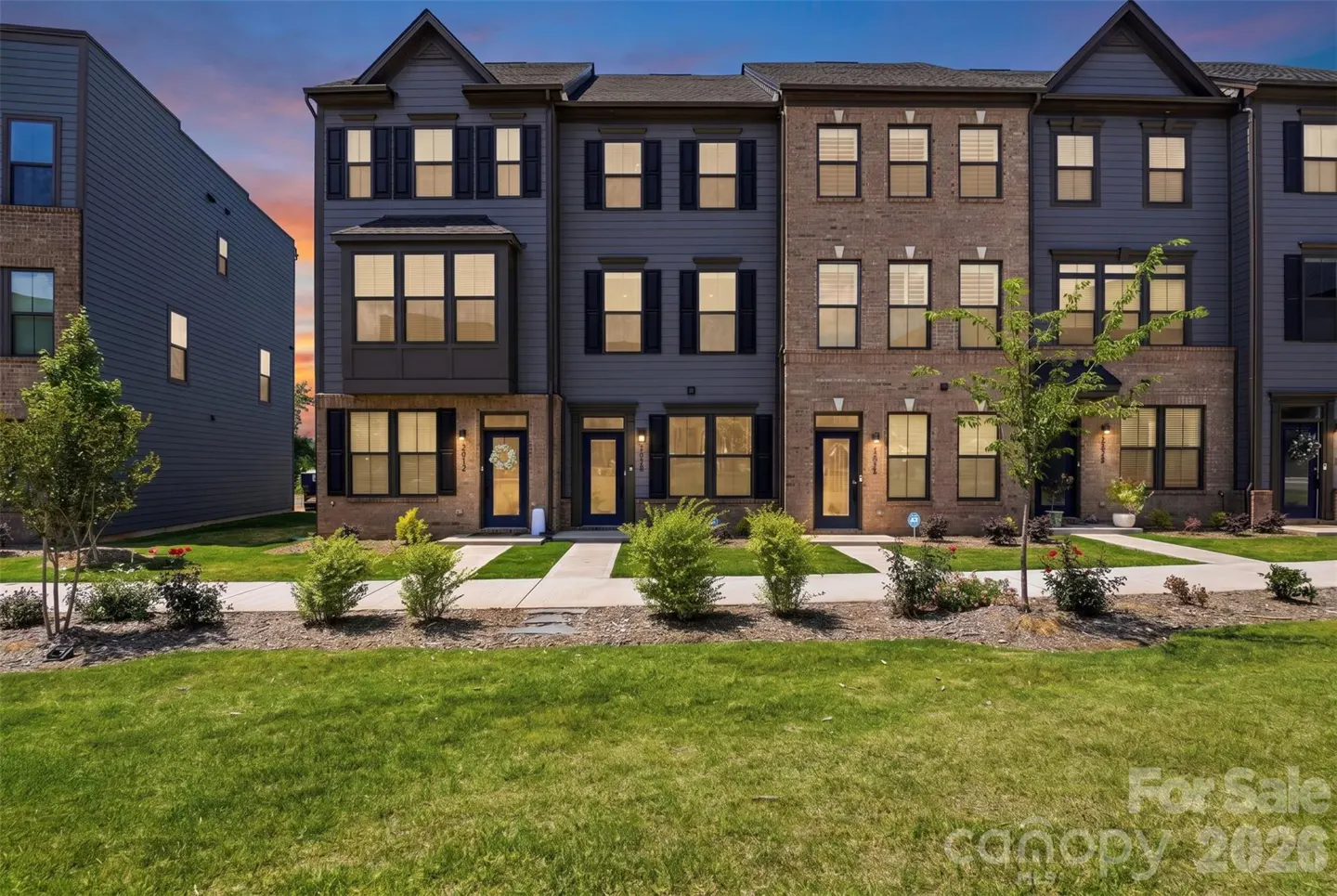 A row of modern three-story townhomes with dark blue siding and brick accents under a sunset sky, with a grassy front lawn.
