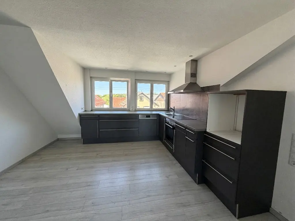 A bright kitchen with gray cabinets, stainless steel appliances, and light wood floors under a slanted ceiling. Windows overlook a neighborhood.