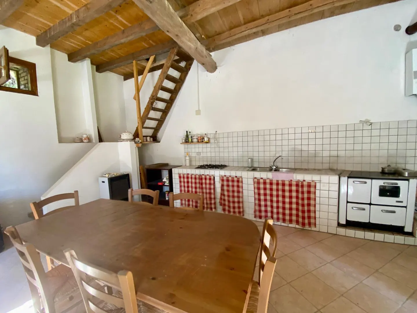 Rustic kitchen with a wooden table and chairs, white walls, tiled backsplash, and a wooden ladder leading to the loft. Red and white checkered curtains.