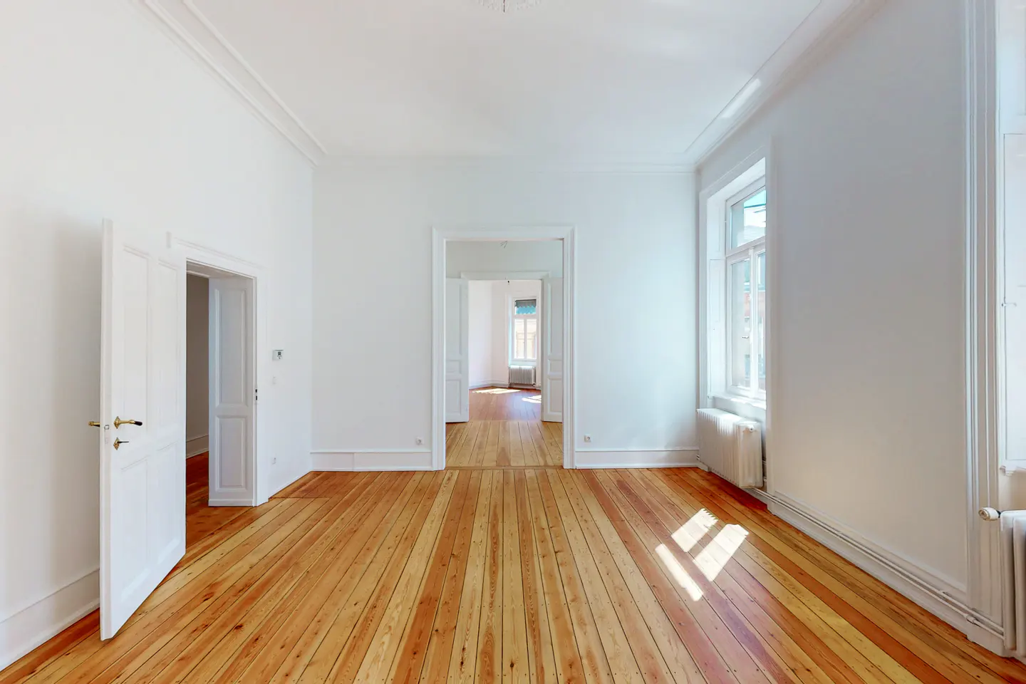 Bright, empty room with wood floors and white walls. An open door leads to another room. A window lets in natural light.