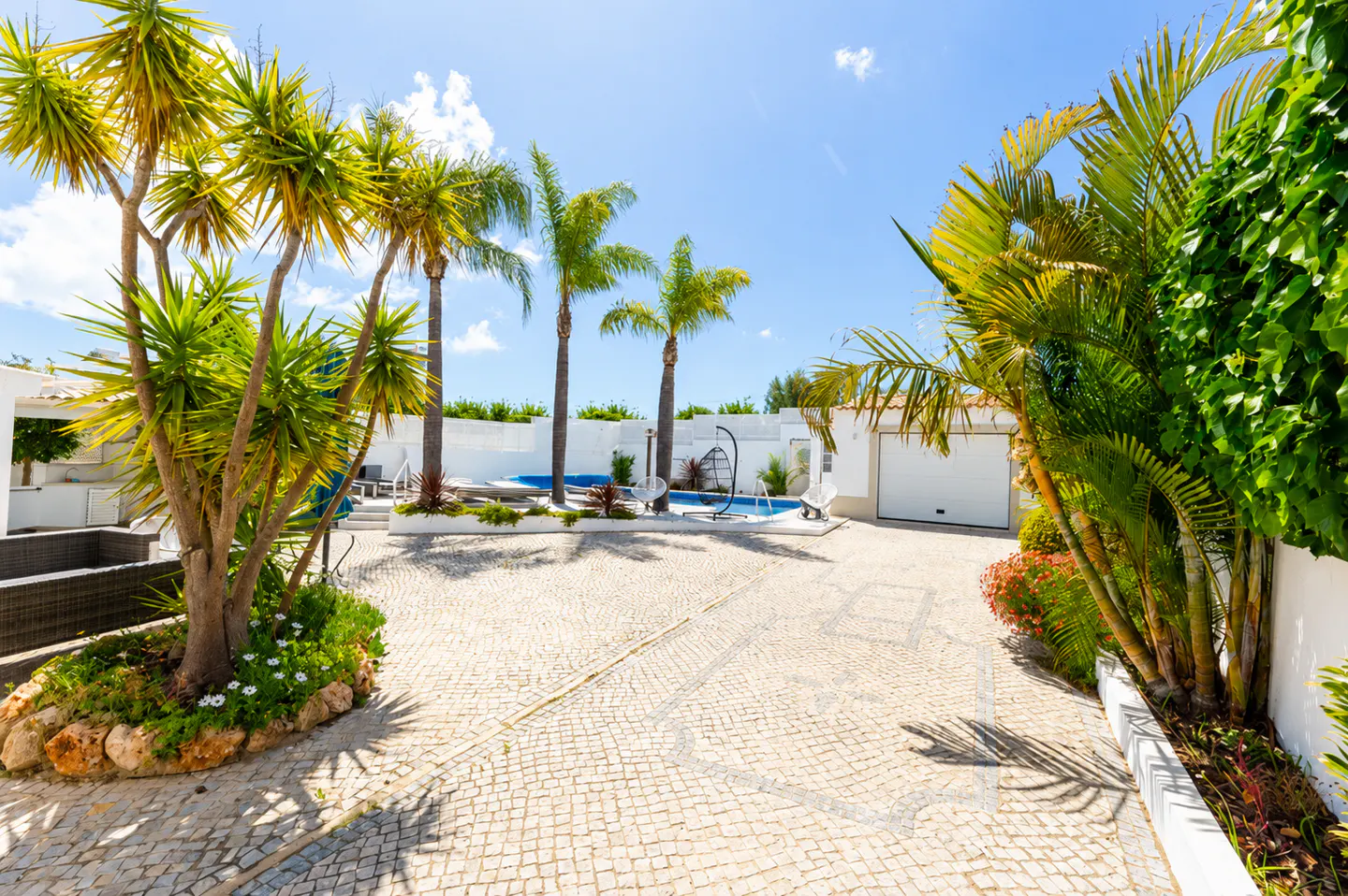 Cobblestone driveway leading to a white house with a pool, palm trees, and lush greenery under a blue sky.
