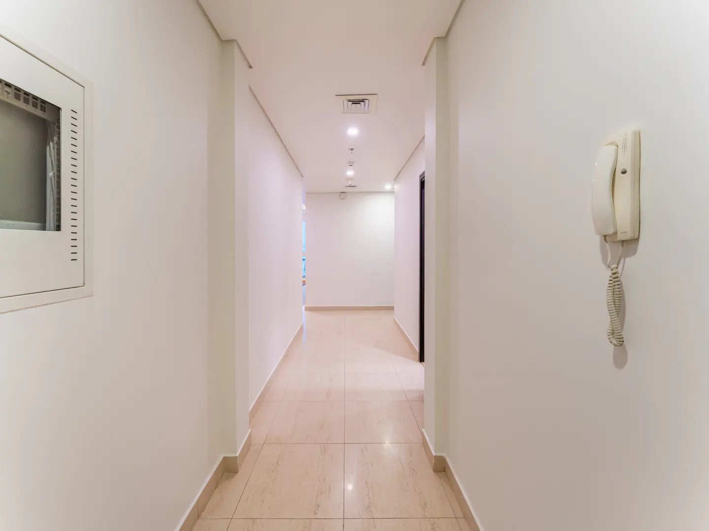 A bright, empty hallway with beige tile floors and white walls. A wall-mounted phone is on the right.