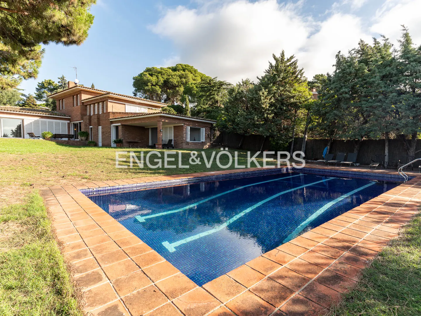 Exterior view of a two-story brick house with a blue tiled pool and green lawn. Engel & Völkers logo is visible.