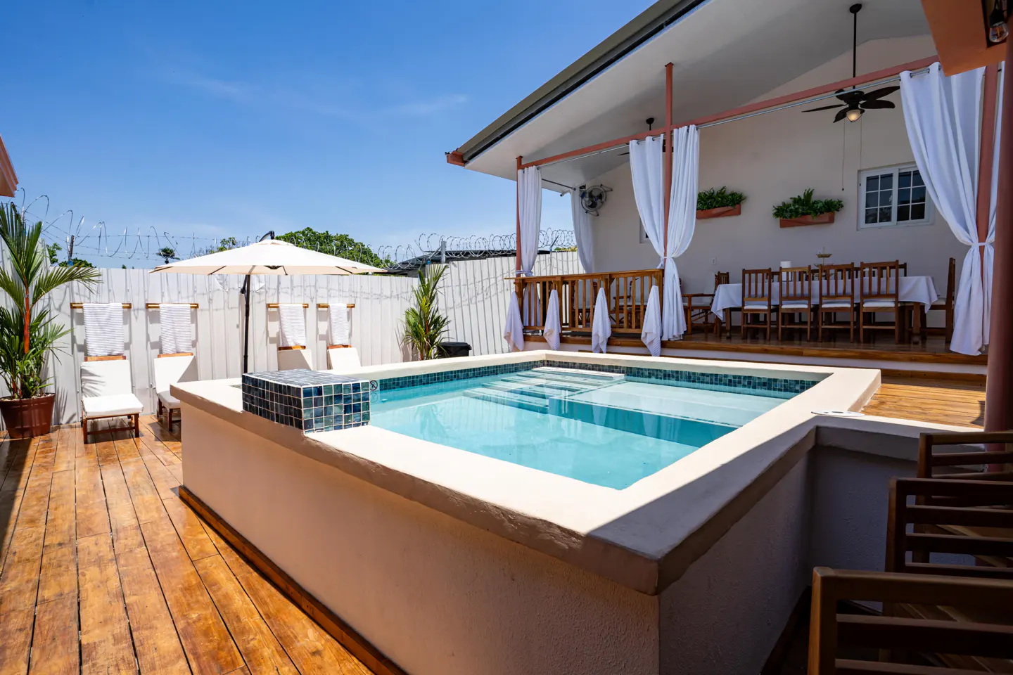 Outdoor pool with blue tiles and clear water, surrounded by wooden deck, lounge chairs, and a covered dining area with white curtains.