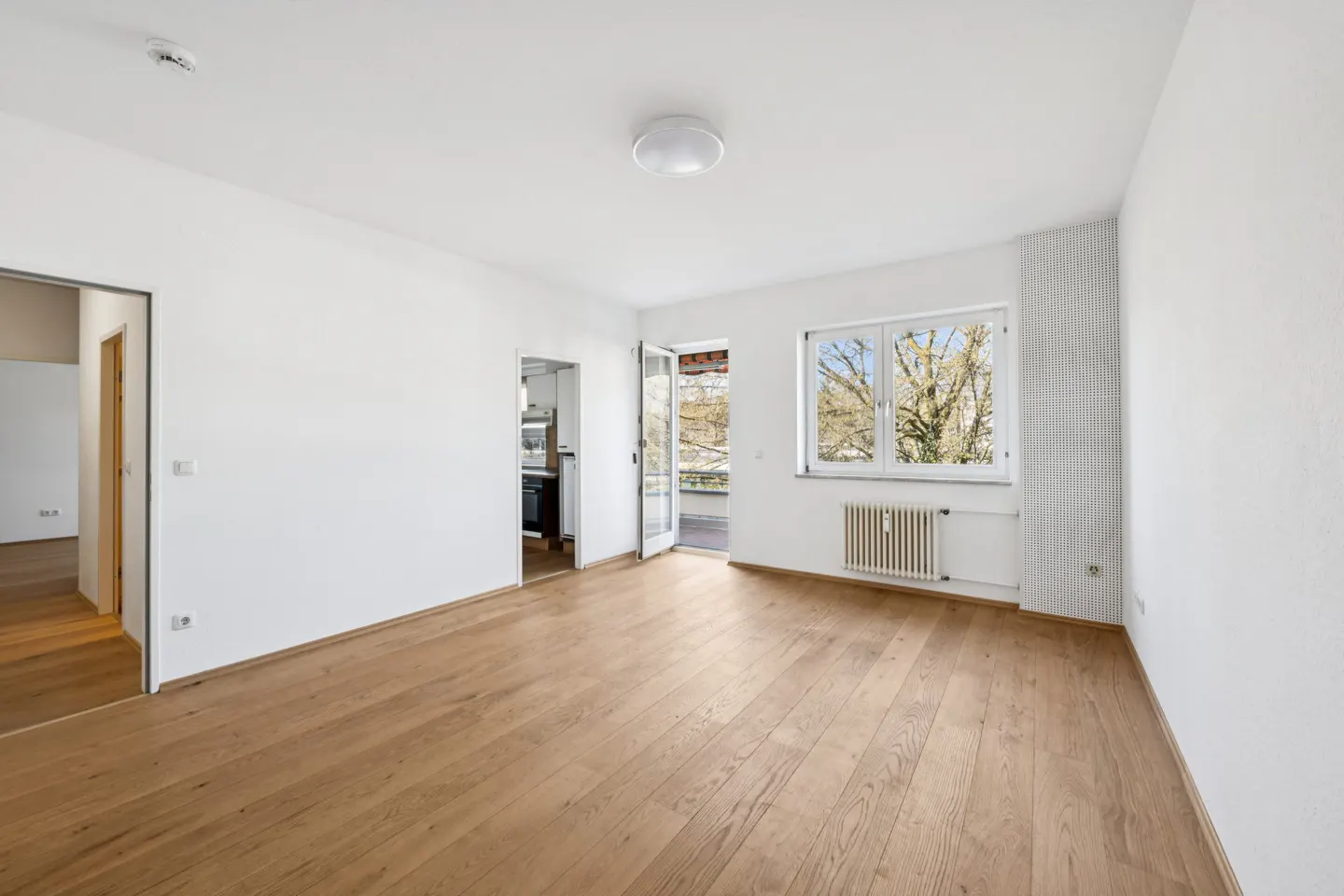 Bright, empty room with wood floors, white walls, and a window. A door leads to a balcony, and another to a kitchen.