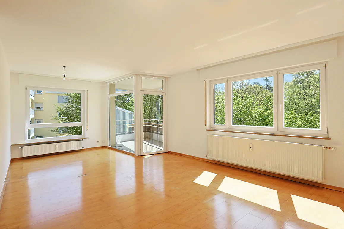 Bright, empty room with wood floors, white walls, and large windows. A balcony is visible through glass doors. Radiators are under the windows.