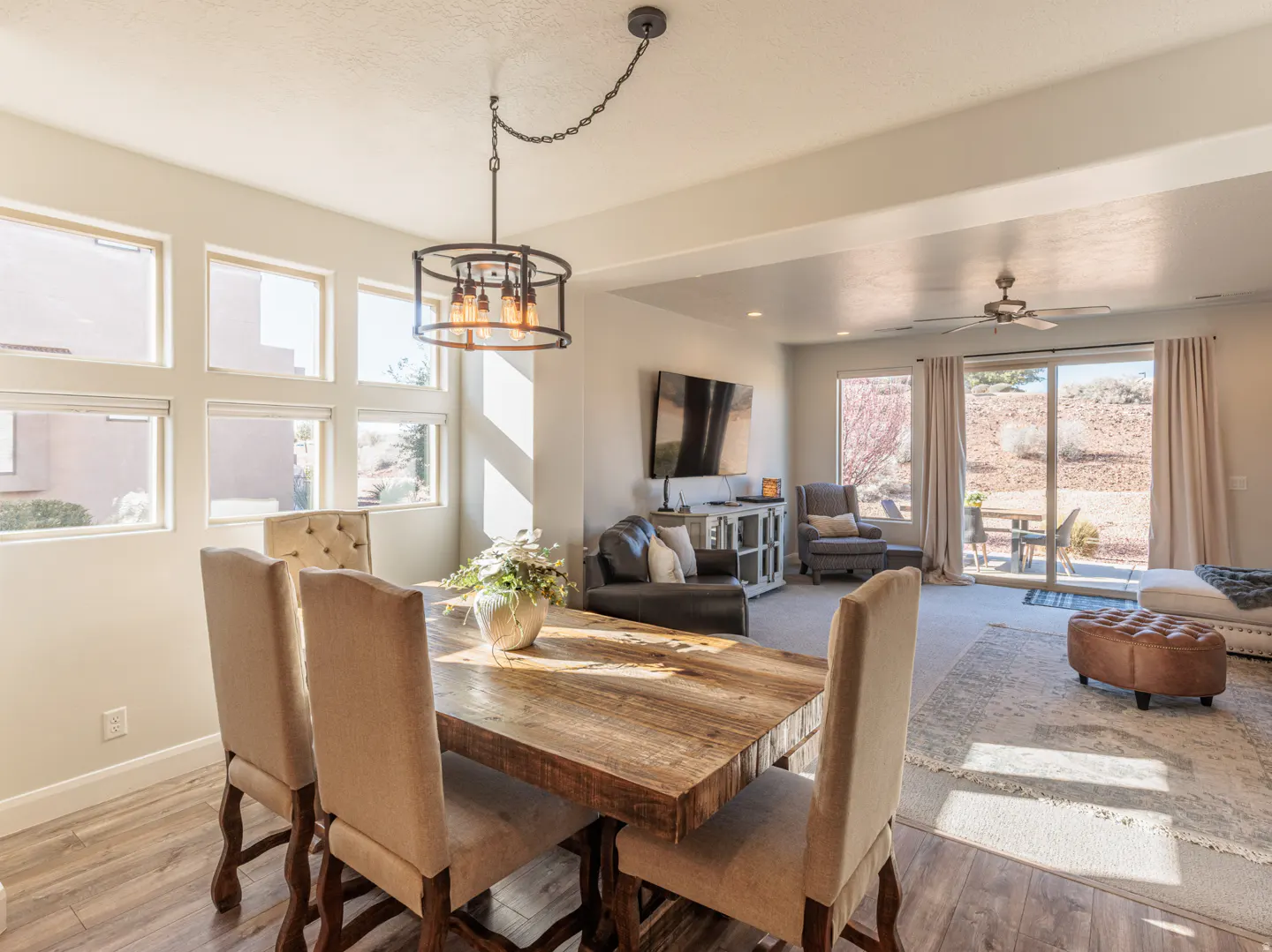 Open-concept dining and living area with a rustic wood table, upholstered chairs, and a view of a desert patio.