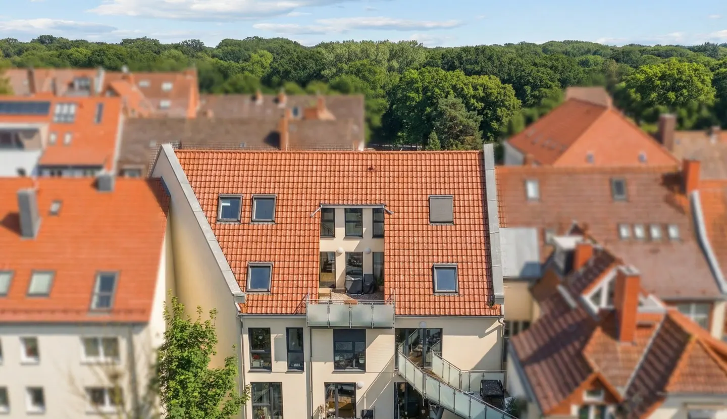 Aerial view of a modern apartment building with a red tile roof, surrounded by older buildings and green trees.