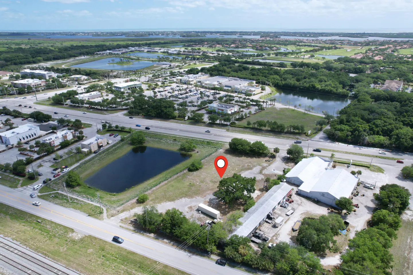 Aerial view of a vacant lot with a red location pin, near a pond, road, and commercial buildings. Trees and greenery surround the area.