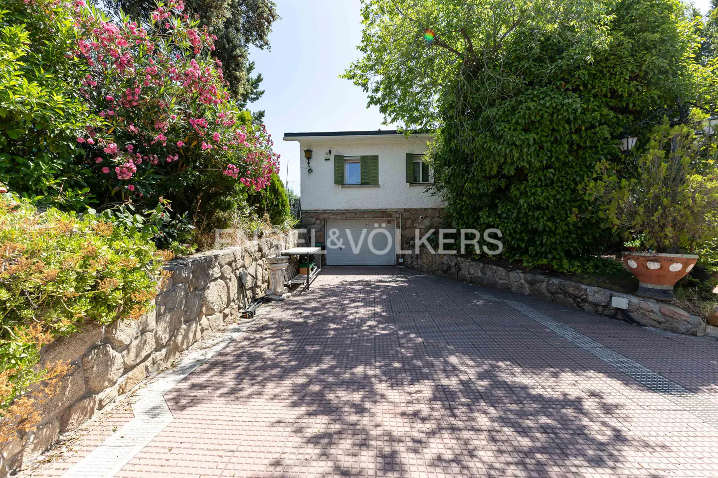 Two-story white house with green shutters and a garage, framed by trees and pink flowers. A brick driveway leads to the house.