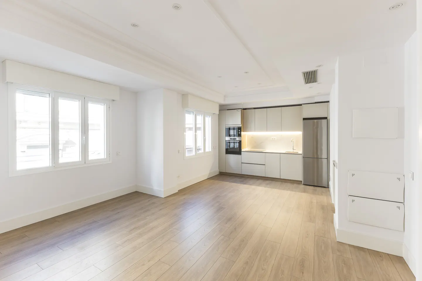 Bright, empty apartment with wood floors, white walls, and a modern kitchen with stainless steel appliances. Natural light streams through the windows.