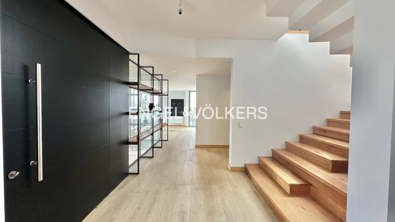 Modern home foyer with a black front door, metal and glass shelving, light wood floors, and a light wood staircase against white walls.