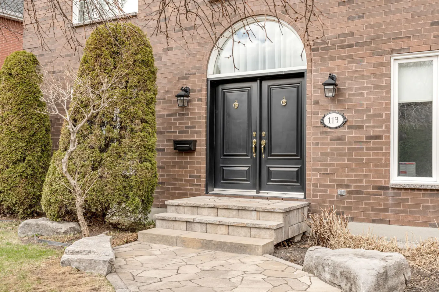 Brick house with black double doors, arched window above, and stone steps. Address number 113 is visible. Tall green bushes flank the entrance.