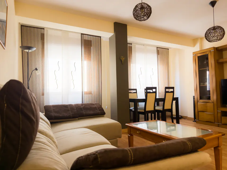Living room with a beige sectional sofa, glass-top coffee table, and a dining area with chairs near large windows.