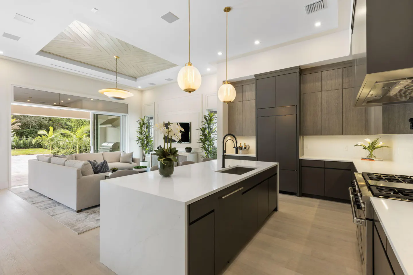 A modern kitchen with a large island, dark cabinets, white countertops, and pendant lights. A living room is visible in the background.