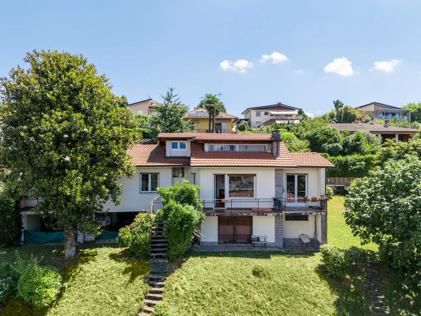 A white two-story house with a red tile roof sits on a green hillside under a blue sky. A large tree is on the left.
