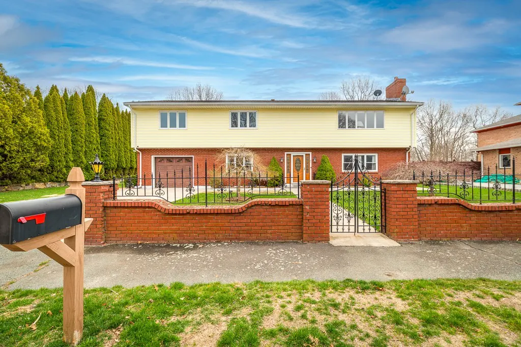 Two-story house with yellow siding and a brick facade, surrounded by a brick wall with a black iron gate.