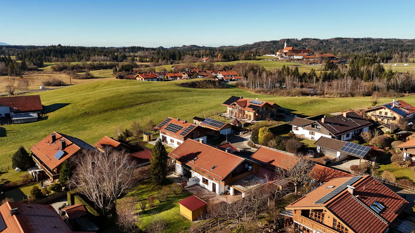 Aerial view of a rural neighborhood with houses with red roofs and solar panels, green fields, and a church in the background.