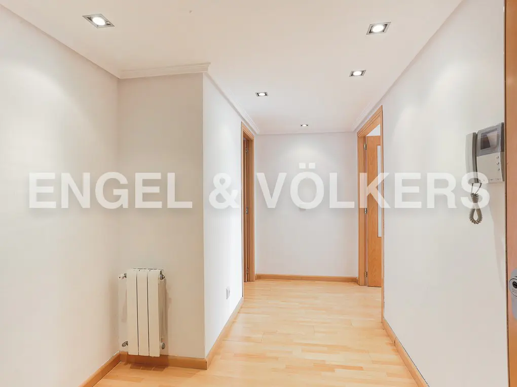 Bright hallway with light wood floors, white walls, recessed ceiling lights, a white radiator, and two wooden doorways.