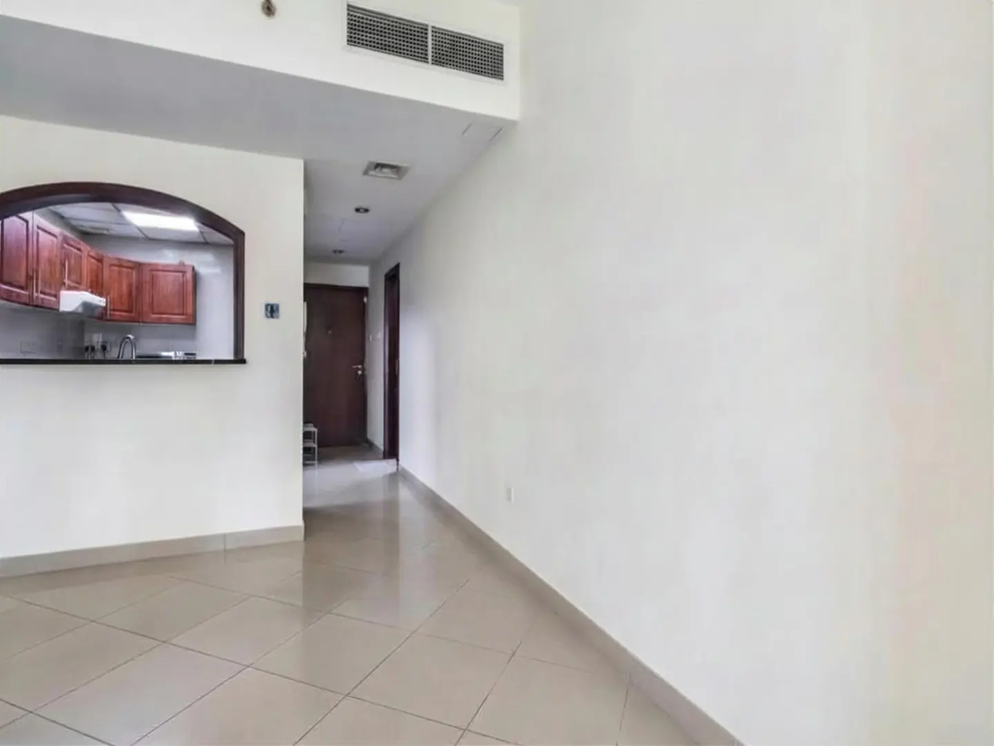 An interior shot of a vacant apartment with white walls and beige tile flooring. A kitchen with wood cabinets is visible through an arched opening.