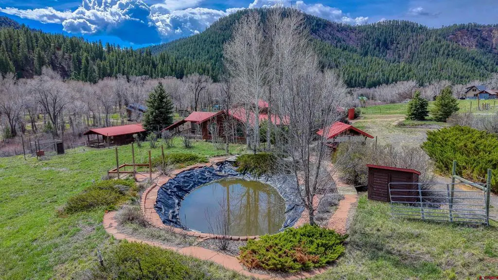 Aerial view of a property with red-roofed buildings, a pond lined with brick, and a backdrop of green mountains and a partly cloudy sky.