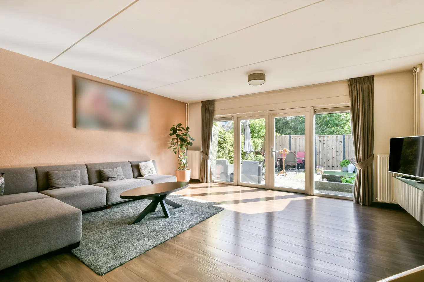 Living room with gray sectional sofa, dark wood floors, and glass doors to a patio with green foliage.