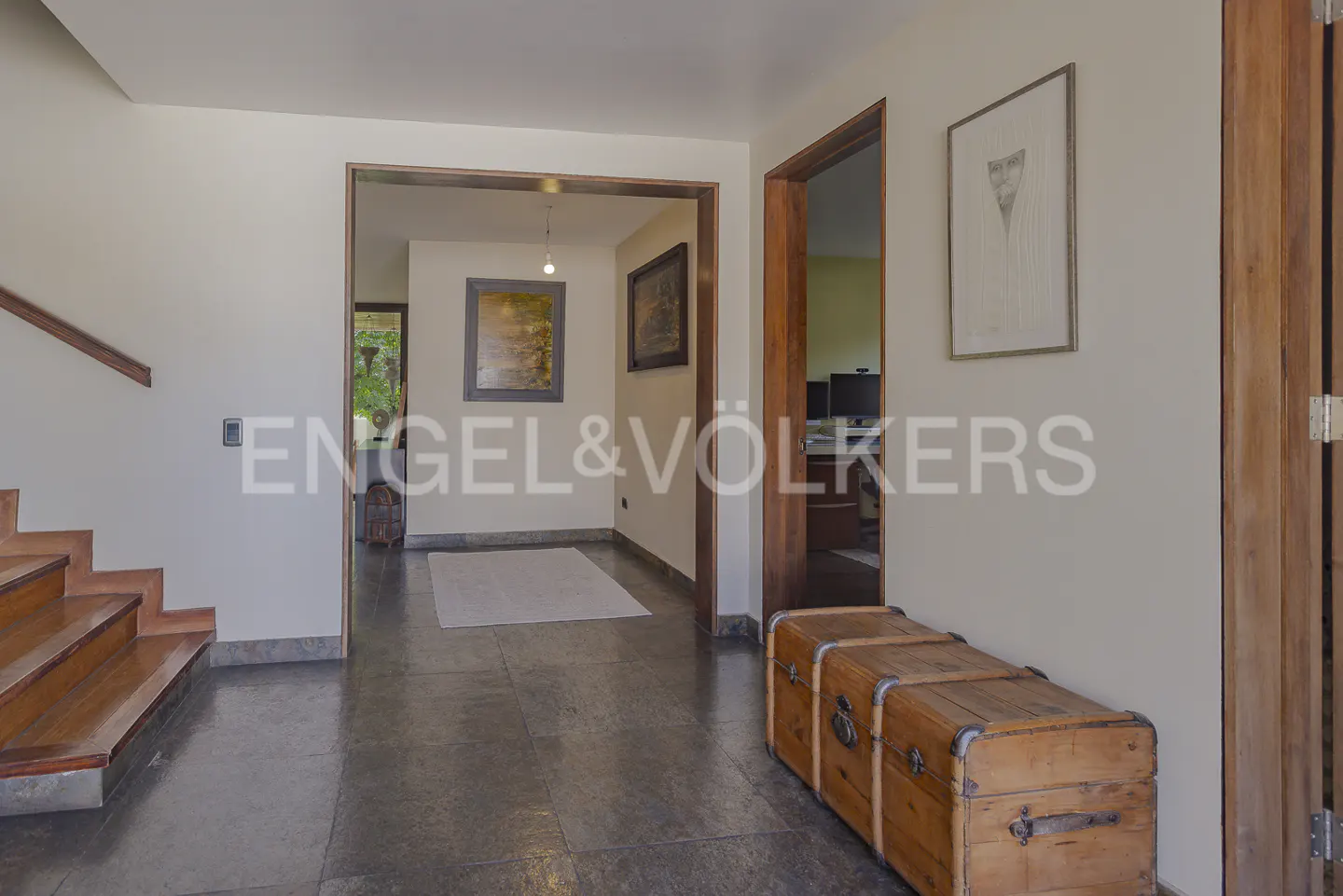 Foyer with wooden stairs, dark tile floor, and two wooden chests. Artwork hangs on the walls.