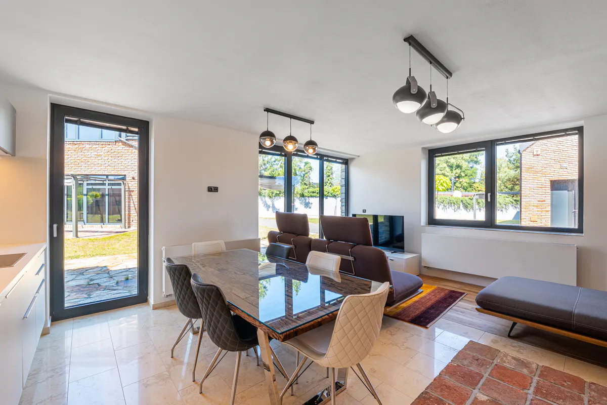 Bright, modern living and dining area with a glass-topped table, brown leather sofa, and large windows overlooking a green yard.