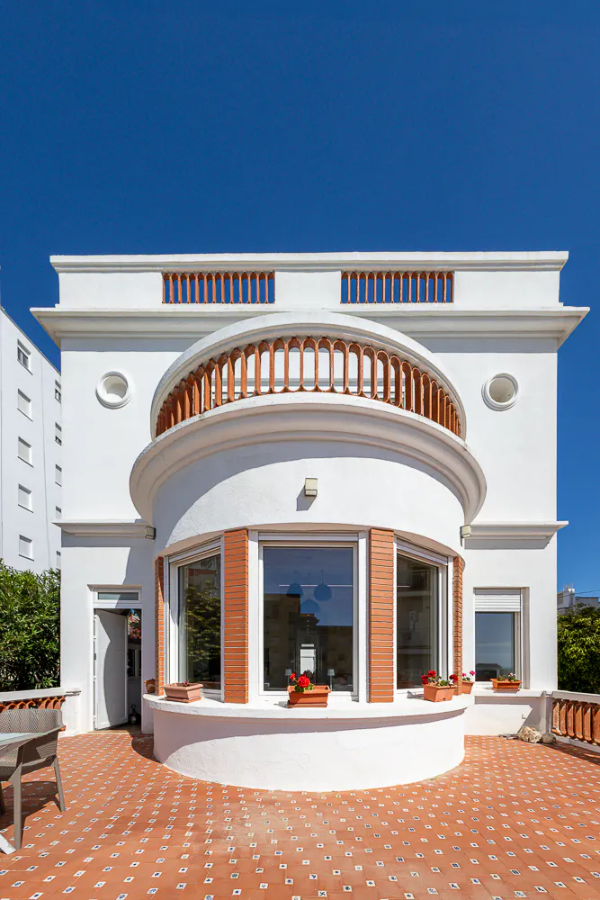 Exterior view of a white, art deco building with a curved balcony and terracotta tile roof under a clear blue sky.