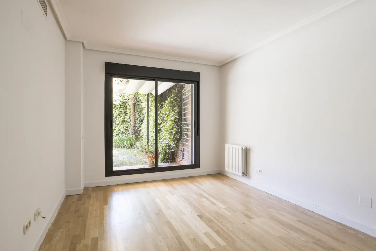 Bright, empty room with light wood floors, white walls, and a black-framed sliding glass door to a green garden.