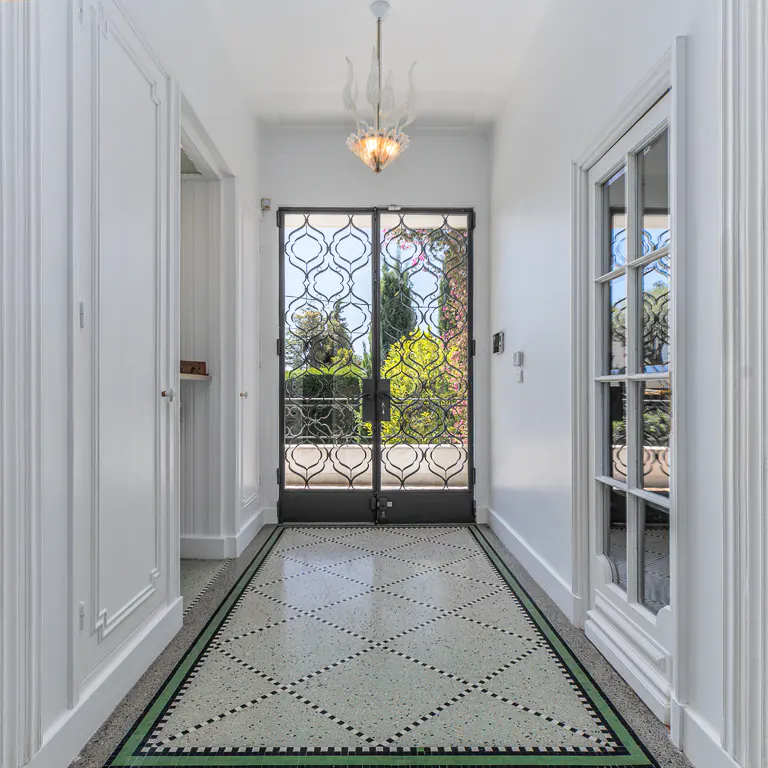 Bright white foyer with a decorative black metal gate to a garden view. A patterned tile floor and a modern chandelier add elegance.