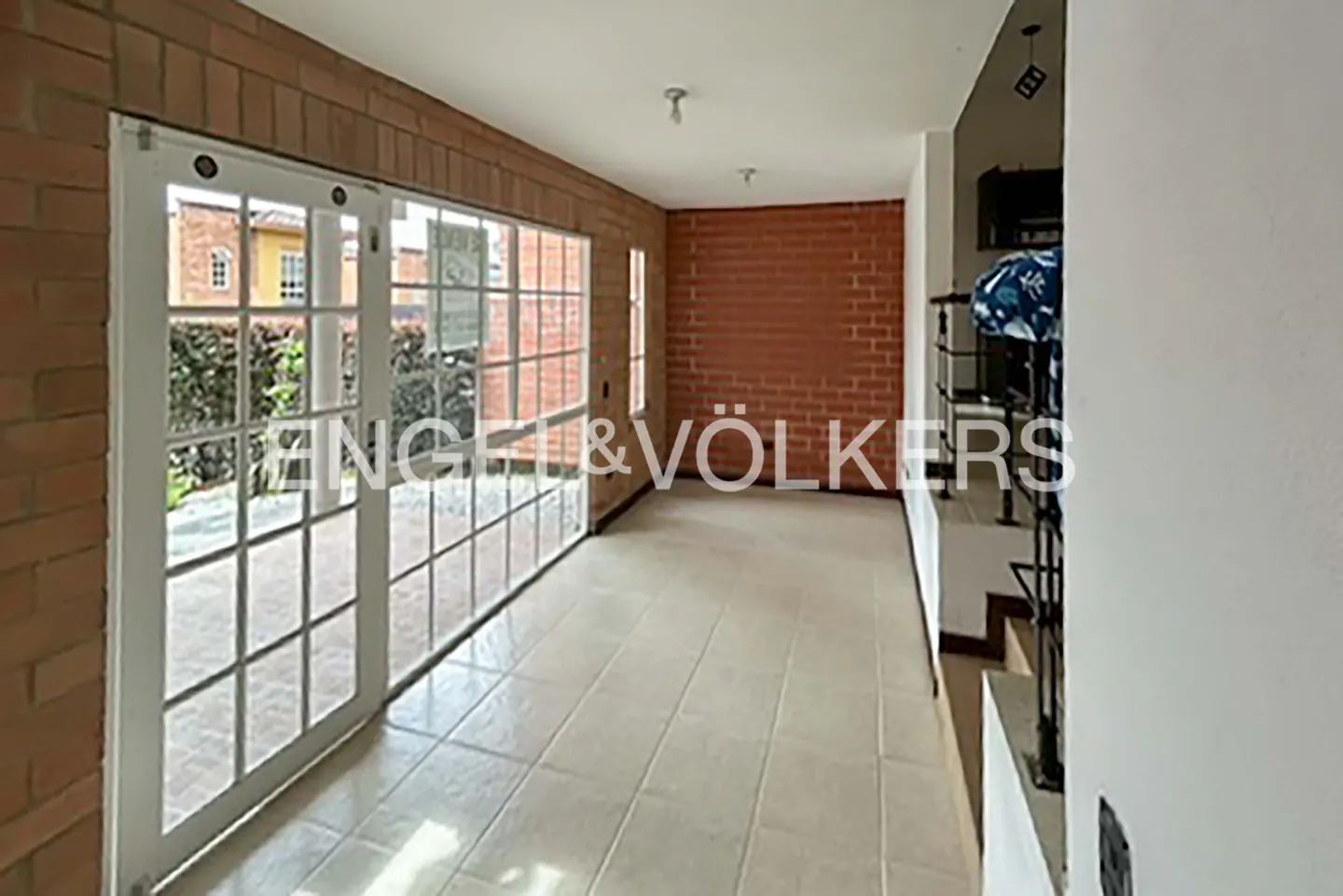 Bright room with brick walls and tile floor. White framed glass doors lead to a garden. Staircase with black railing is visible.