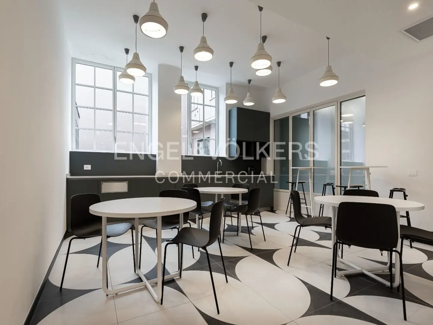 A bright break room with white walls, black chairs, and round white tables. Pendant lights hang from the ceiling. The floor has a black and white pattern.