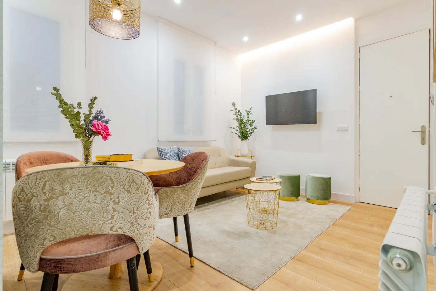 Bright living room with a round table, upholstered chairs, a beige sofa, and a wall-mounted TV. A gold chandelier hangs above the table.