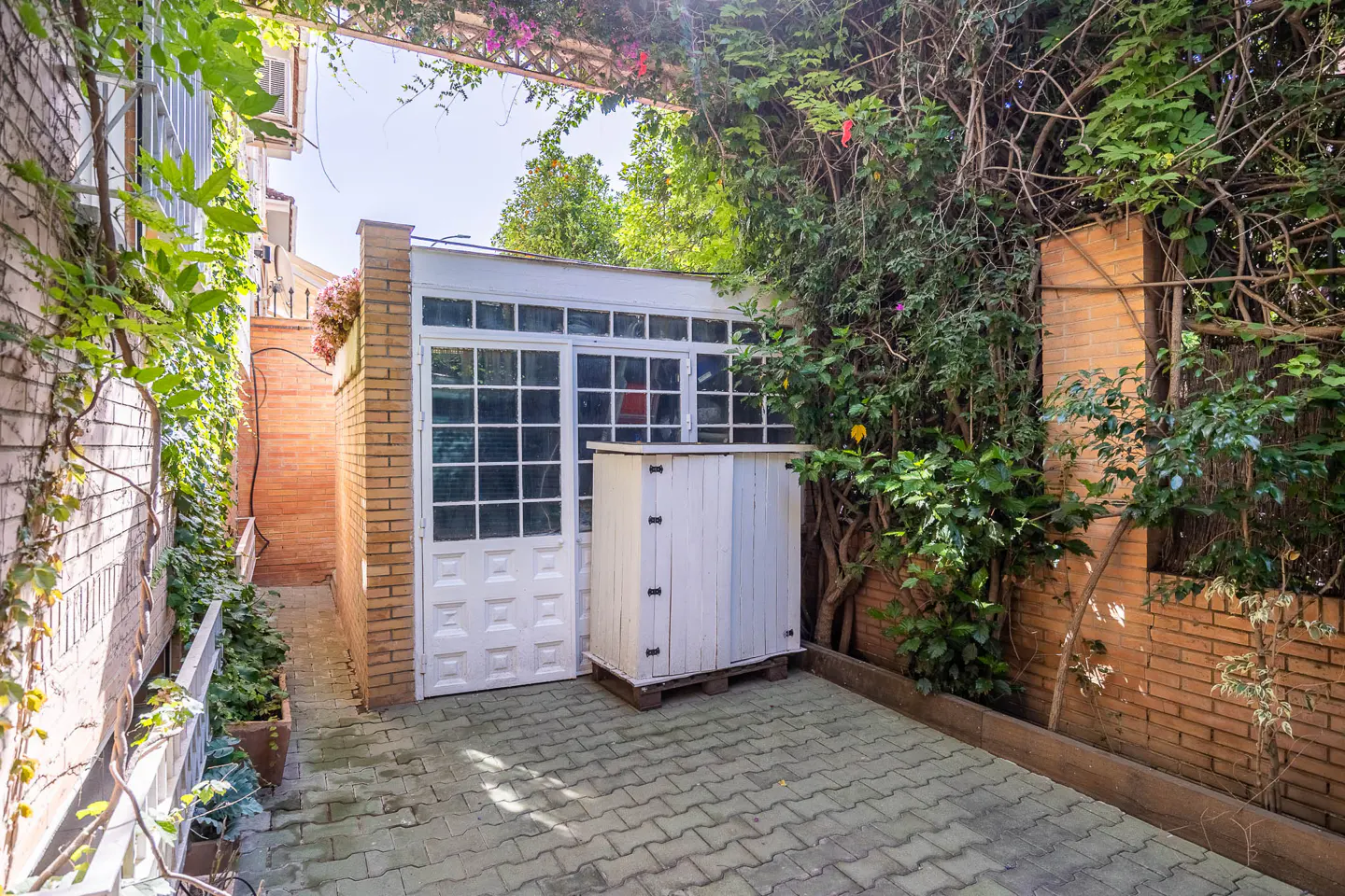 A brick-paved courtyard with a white door and a storage box, surrounded by brick walls and lush greenery.