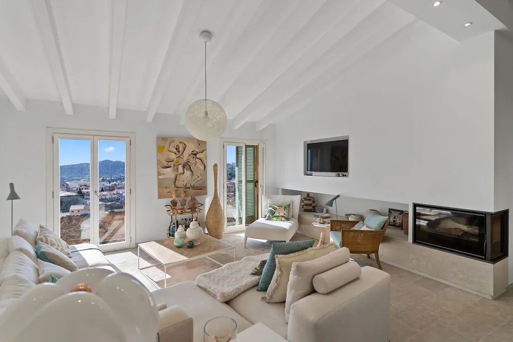 Bright living room with white walls, beamed ceiling, and a large sectional sofa. A fireplace, TV, and artwork add to the room's ambiance.
