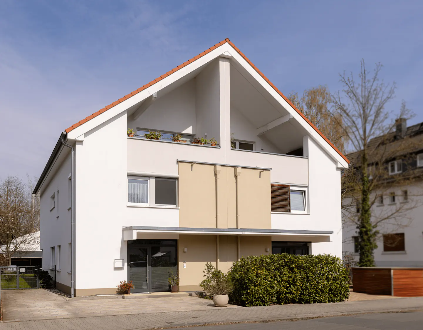 A modern two-story white house with a red tile roof and a balcony with flowers. A green bush is in front.