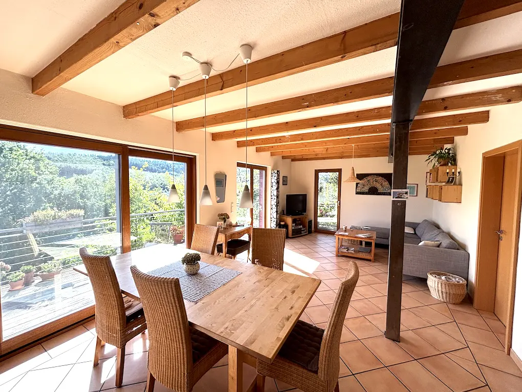 Bright, open-plan dining and living area with wooden beams, tile floor, and large windows overlooking greenery. Dining table with wicker chairs in foreground.