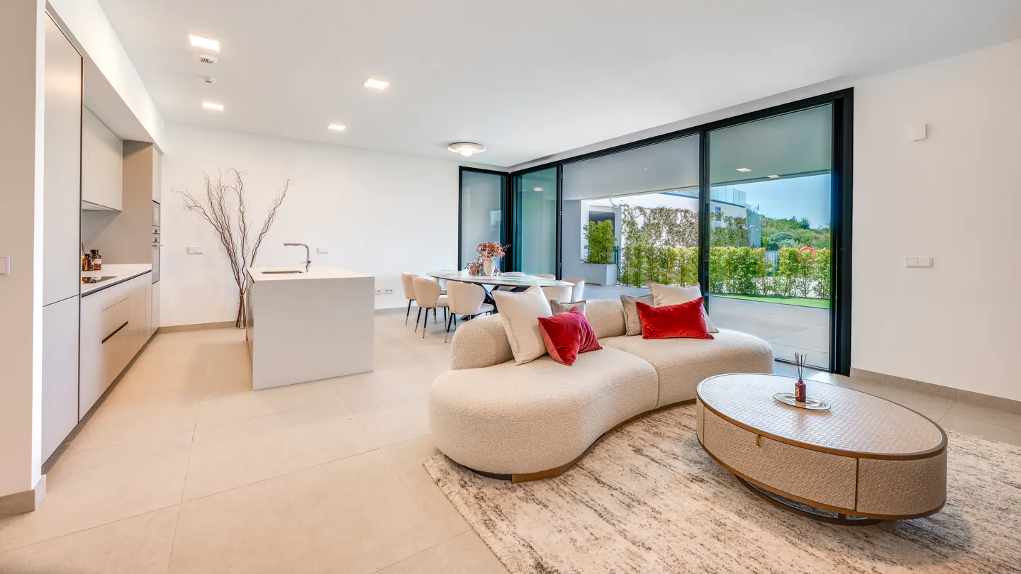 Bright, modern living space with a curved sofa, red pillows, and a view of the outdoors through large glass doors.
