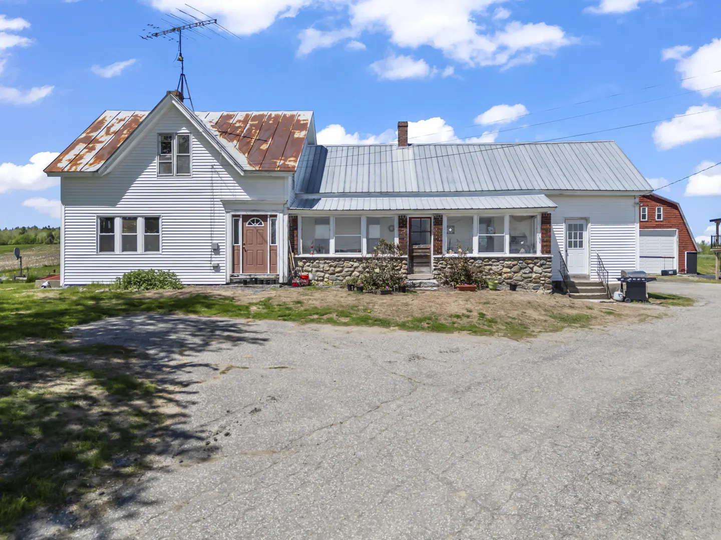 Exterior view of a white farmhouse with a rusty metal roof and a stone foundation under a bright blue sky.