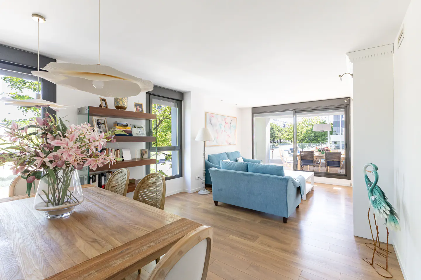 Bright, modern living room with wood floors, blue sofa, dining table with flowers, and balcony view.