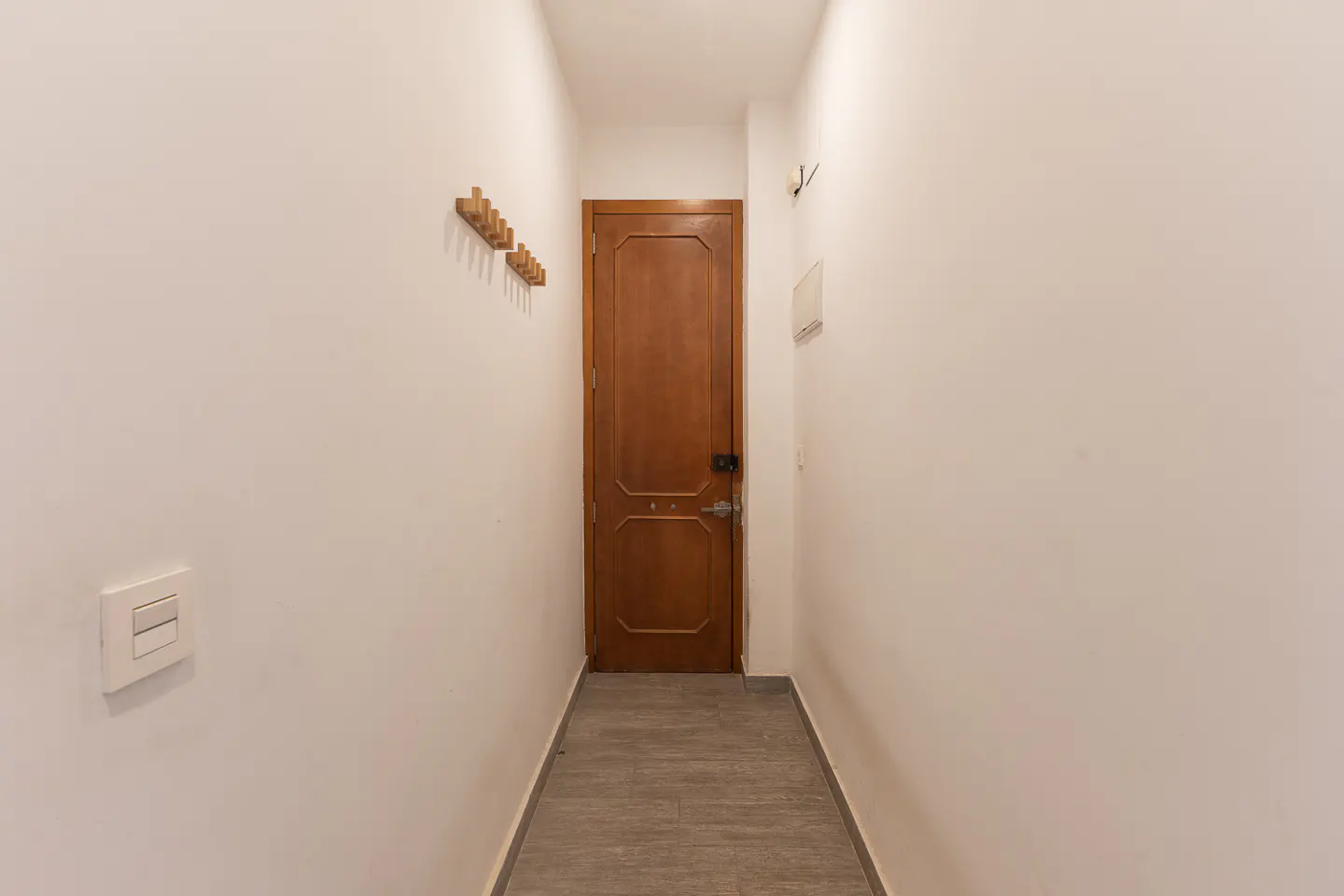 Narrow hallway with white walls, grey tiled floor, a wooden door at the end, and a wall-mounted coat rack on the left.