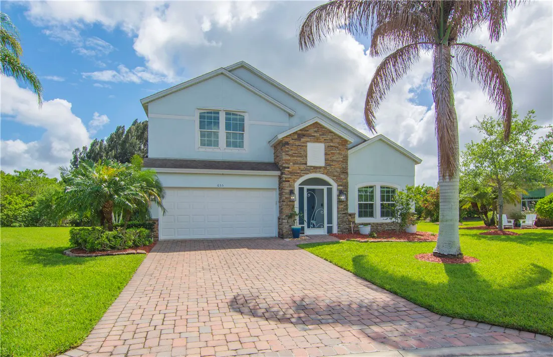 Two-story light blue house with a stone facade, white garage door, and brick driveway. Palm trees and green lawn surround the property.