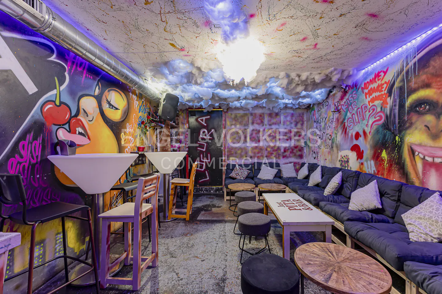 Interior of a bar with graffiti art on the walls, cloud-like ceiling decor, and blue cushioned seating. Tables and chairs are arranged for customers.