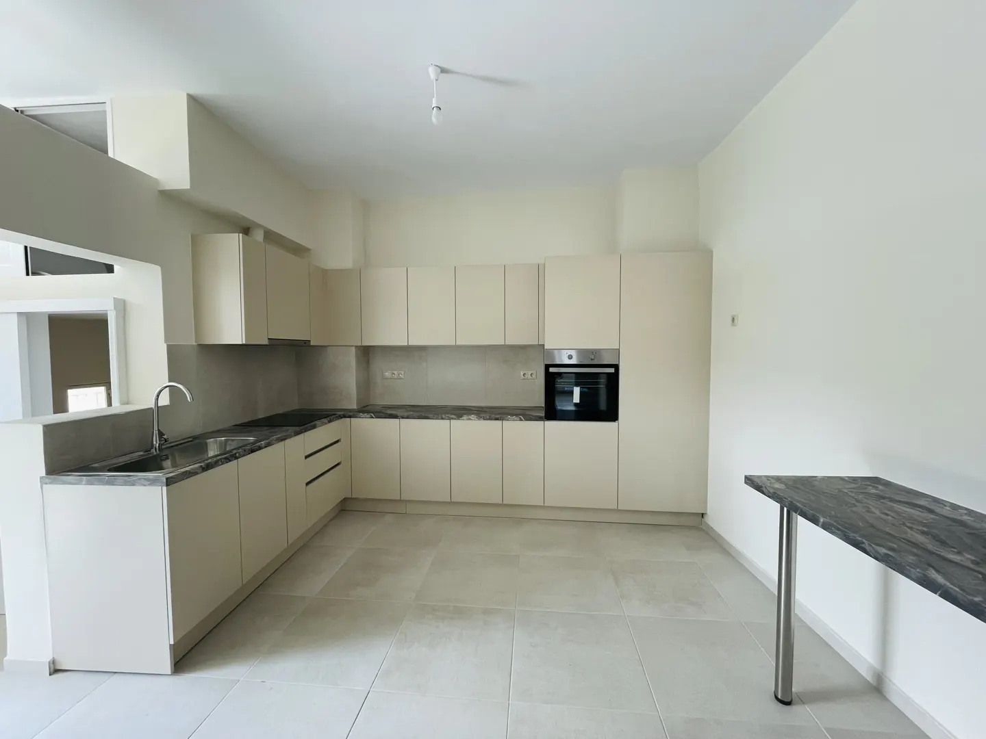 Bright kitchen with beige cabinets, gray countertops, and tile flooring. A stainless steel sink and black oven are visible. A dark marble table stands against the wall.