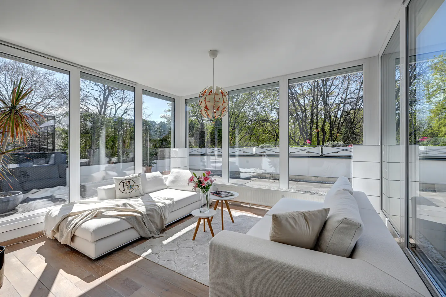 Bright sunroom with white sofas, two small round tables, a patterned rug, and floor-to-ceiling windows overlooking trees.